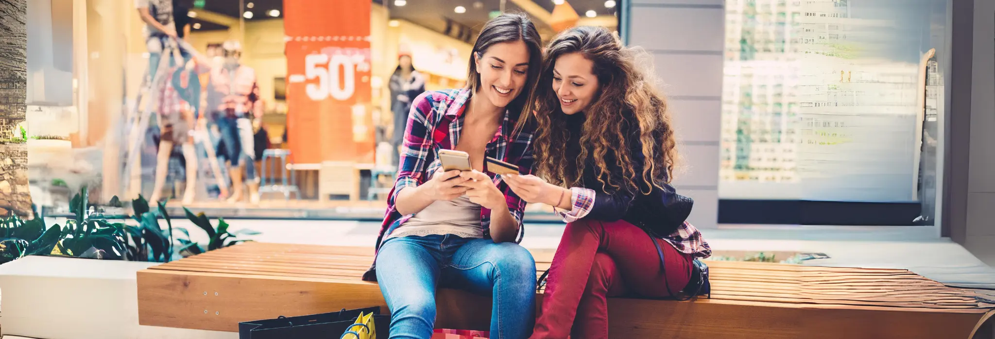 Δύο κοπέλες σε εμπορικό κέντρο κρατούν κάρτα και κινητό.