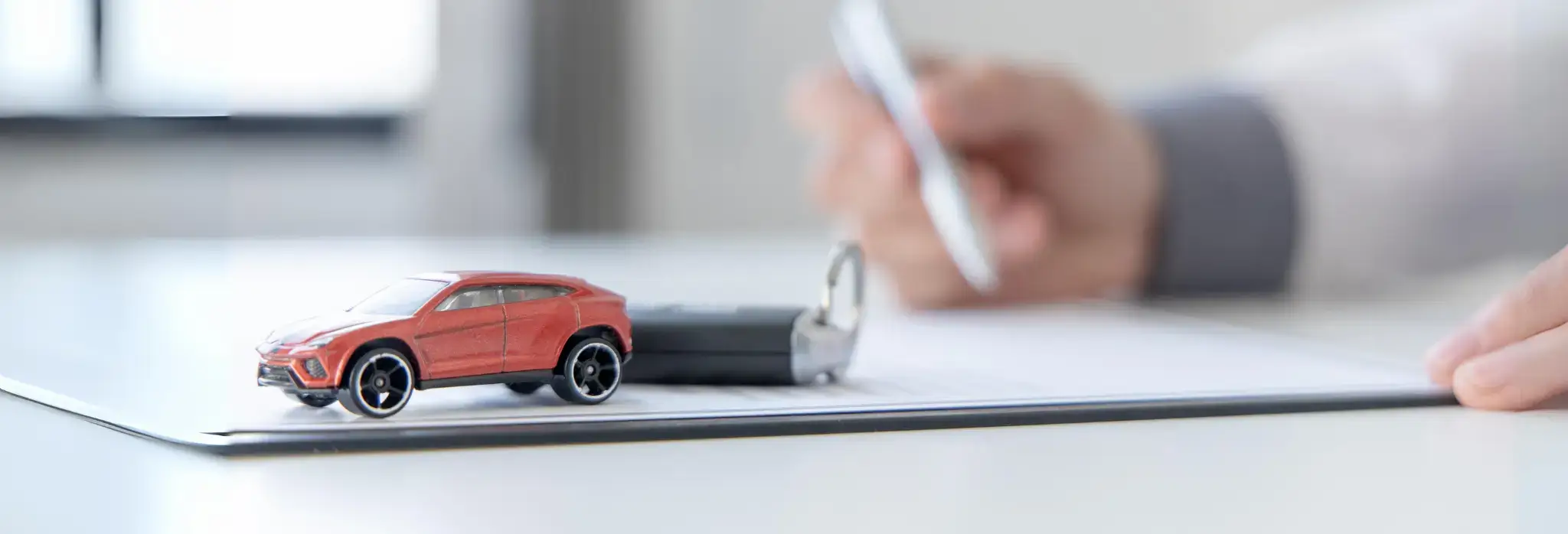 Χέρι με στυλό υπογράφει συμβόλαιο πάνω στο οποίο έχει κλειδιά αυτοκινήτου και μινιατούρα αυτοκινητάκι.