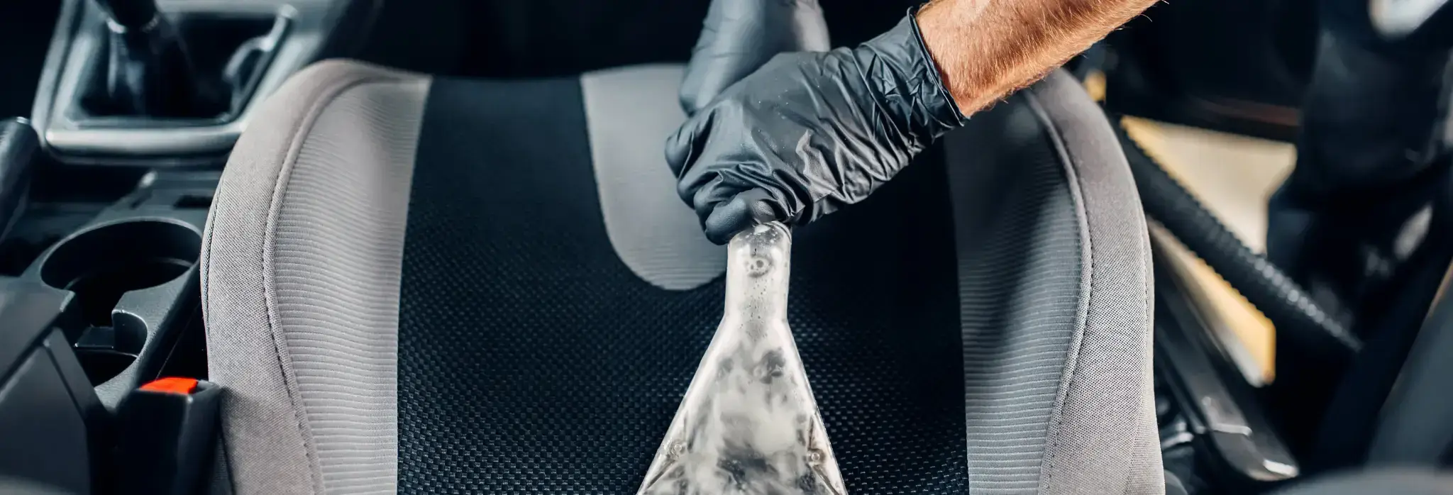 Χέρι με μαύρο γάντι χρησιμοποιεί απορροφητική συσκευή για βαθύ καθαρισμό υφασμάτινου καθίσματος αυτοκινήτου, αφαιρώντας λεκέδες.
