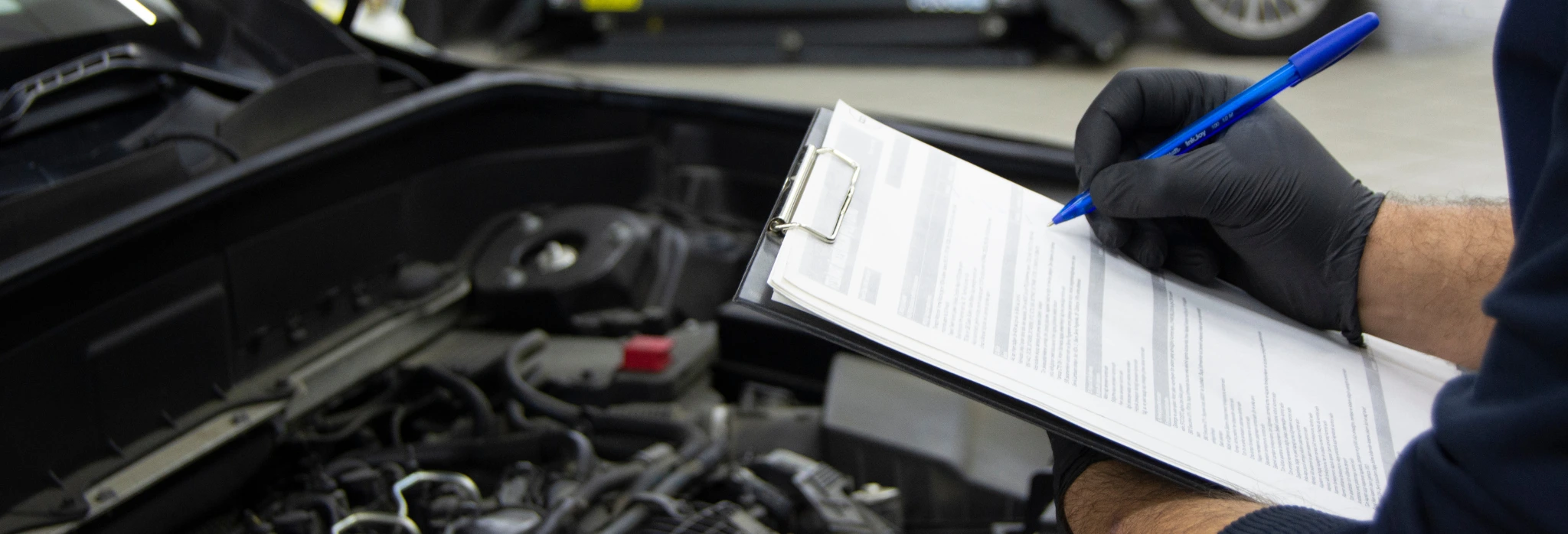 Τεχνικός με μαύρα γάντια καταγράφει στοιχεία σε έντυπο ελέγχου μπροστά από ανοιχτό καπό αυτοκινήτου σε συνεργείο.