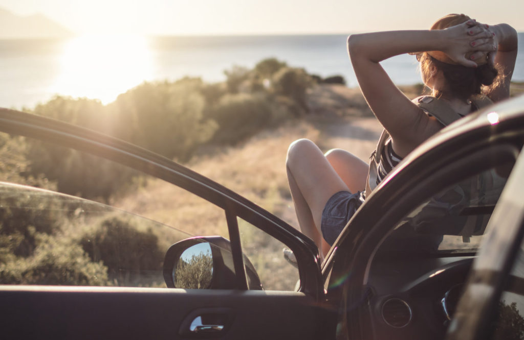 Κοπέλα κάθεται στο καπό του αυτοκινήτου και κοιτάει το ηλιοβασίλεμα στη θάλασσα.